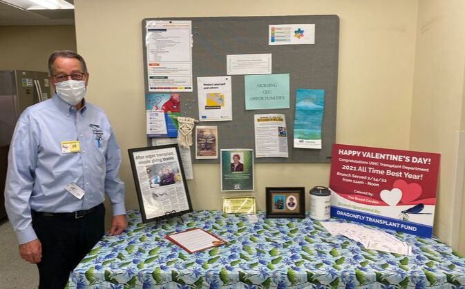 Man in button and medical mask stands next to table with dragonfly table covering and sign displaying information about the Dragonfly Transplant Fund